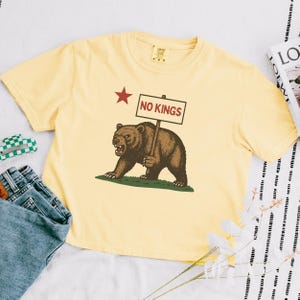 Yellow Comfort Colors cropped t-shirt with NO KINGS bear protest design, styled with book and accessories.