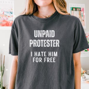 Model wearing vintage black tee with protest slogan “Unpaid Protester – I Hate Him For Free” styled with soft curls and layered necklace.