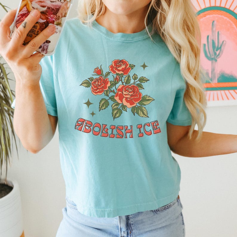 Aqua blue cropped tee with vintage red rose artwork and Abolish ICE slogan, woman taking selfie