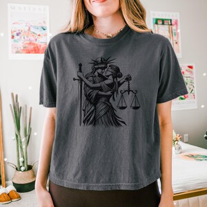 Dark gray cropped Comfort Colors tee with black line art of Statue of Liberty embracing Lady Justice, modeled in a modern bedroom