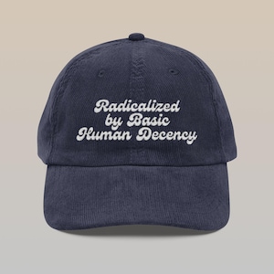 Op de afbeelding: Marineblauwe corduroy baseballcap met de geborduurde tekst "Radicalized by Basic Human Decency" in witte letters. De cap heeft een gebogen klep en een knoop aan de bovenkant.