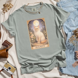 May include: A light green t-shirt featuring a graphic of a Siamese cat under a full moon, surrounded by flowers and trees. The word "SIAMITY" is printed below the image. The shirt is made of a soft material.