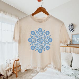May include: Cream-coloured t-shirt with a light blue floral design. The design is symmetrical, featuring a central pattern of leaves and flowers. The shirt is hanging on a wooden hanger.