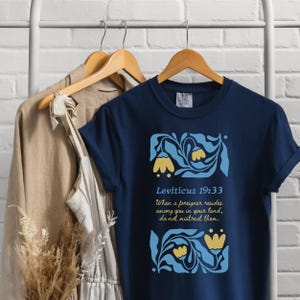 Navy Shirt on Hanger with Linen Dress:
Navy Comfort Colors tee with yellow flower and blue foliage graphic, featuring Bible verse Leviticus 19:33, styled on a hanger display
