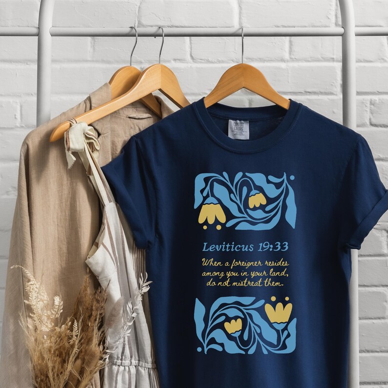 Navy Shirt on Hanger with Linen Dress:
Navy Comfort Colors tee with yellow flower and blue foliage graphic, featuring Bible verse Leviticus 19:33, styled on a hanger display