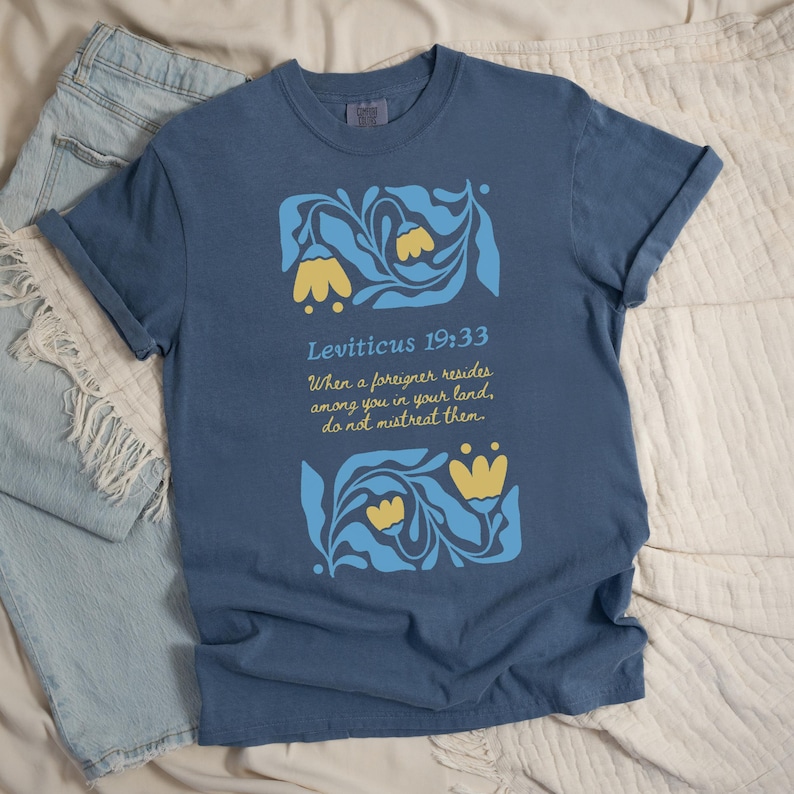 Denim Blue Shirt Flat Lay:
Denim blue Bible verse T-shirt featuring Leviticus 19:33 and vintage folk flowers, laid out with a book and coffee