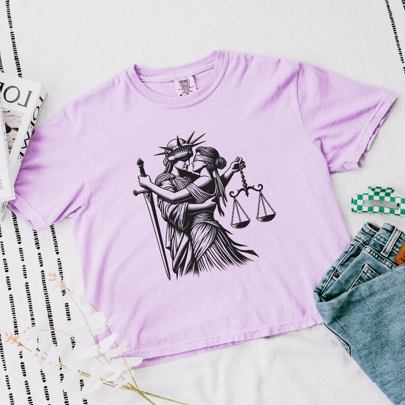 Lilac flat lay t-shirt with sword-wielding Statue of Liberty embracing blindfolded Lady Justice, styled with magazine and jeans