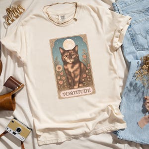 May include: Cream-colored t-shirt featuring a graphic of a cat with the word "TORTITUDE" below. The cat is brown and tan, set against a backdrop of flowers, a full moon, and a blue sky. The shirt is paired with blue jeans.