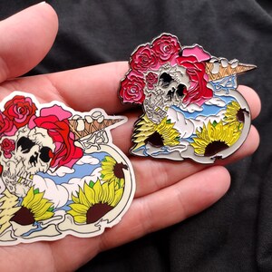 May include: Two skull-shaped enamel pins. One is a sticker, the other is a pin. Both feature a skull with red roses, a pink rose, a blue sky, yellow sunflowers, and a waffle cone. The sticker is on the left, and the pin is on the right.