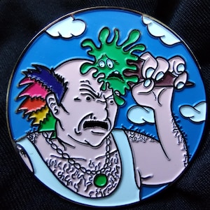 May include: A round enamel pin featuring a cartoon character with rainbow hair, a green goo monster on his head, and a mustache. The character is wearing a white tank top with a gold chain and a green gemstone pendant. The background is blue with white clouds.