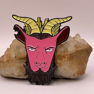 May include: A pink and black enamel pin featuring a stylized goat head with yellow horns and a black beard. The goat has an angry expression. The pin is set against a neutral background.