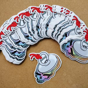 May include: A stack of colorful skull-shaped stickers with a red string attached to each one. The stickers are arranged in a semi-circle with one sticker at the bottom of the image.