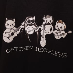 May include: Black t-shirt with a white graphic of four cats playing musical instruments. The cats are wearing hats and have different expressions. The text "Catchen Meowlers" is printed below the cats.