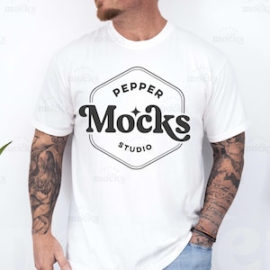 May include: A white t-shirt with a black logo that says "Pepper Mocks Studio" in a hexagon shape. The person wearing the shirt has tattoos on their arms.