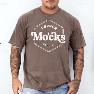 Puede incluir: Una camiseta marrón con un logotipo blanco que dice "Pepper Mocks Studio". La persona que lleva la camiseta tiene tatuajes en los brazos.