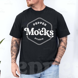 Op de afbeelding: Een zwart T-shirt met een wit logo met de tekst "Pepper Mocks Studio". De persoon die het shirt draagt heeft tatoeages op zijn armen.