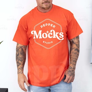 Puede incluir: Una camiseta naranja brillante con un logotipo blanco que dice "Pepper Mocks Studio" en una fuente retro.