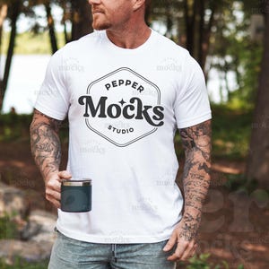 Può includere: Una maglietta bianca con un logo in bianco e nero che dice "Pepper Mocks Studio". La persona che indossa la maglietta ha dei tatuaggi sulle braccia.