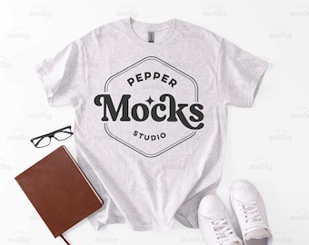 May include: A light gray t-shirt with the text "PEPPER Mocks STUDIO" in a black, retro-style design. The image also includes a brown notebook, black glasses, and white sneakers, all arranged on a white surface.