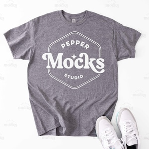 May include: A heather gray t-shirt with a white graphic design that says "Pepper Mocks Studio". The shirt is laid flat on a white surface with two white sneakers beside it.