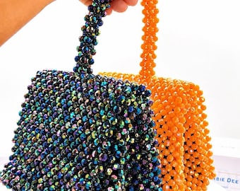 May include: Two structured handbags crafted from faceted beads. One bag features iridescent dark blue and purple beads, while the other is made of bright orange beads. Each bag has a single top handle and a small rectangular gold-toned metal tag.