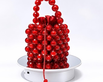 Puede incluir: Un bolso de mano de color rojo brillante construido con cuentas esféricas facetadas y translúcidas. El bolso cuenta con un asa redondeada hecha de las mismas cuentas rojas y un cierre de cordón de satén rojo, con un cordón rojo colgando al frente.