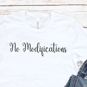 May include: White t-shirt with the text "No Modifications" in black script, paired with rolled-up blue jeans and black canvas sneakers. The items are arranged on a light wood-grain surface.