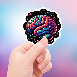 May include: A colorful sticker featuring a stylized brain illustration. The brain is depicted in vibrant shades of blue, pink, and yellow, with a black scalloped border adorned with stars and planets. The sticker is held in a hand.