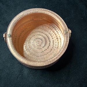 May include: A hammered copper bowl with a spiral pattern on the interior base. The bowl has a rounded shape with two small handles on either side. The copper has a warm, reddish-brown hue.