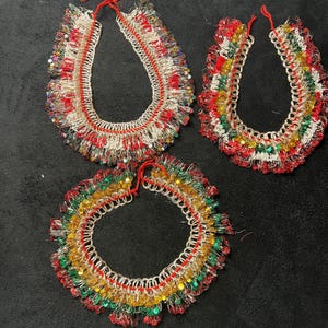 May include: Three handmade circular collars crafted from silver-toned safety pins and vibrant red, green, and yellow beads. Each piece features a flexible structure held together by red thread, creating a textured, layered appearance with metallic accents.