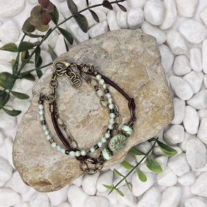 May include: A layered bracelet with brown leather, antique brass chains, and light blue beads. The bracelet includes decorative light blue and green glass beads. The jewellery is displayed on a stone with white pebbles in the background.
