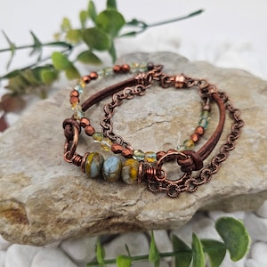 Puede incluir: Un brazalete de capas con un cordón de cuero marrón, una cadena de cobre y cuentas de vidrio en tonos de verde, amarillo y azul. El brazalete tiene un cierre de cobre y un anillo de cobre decorativo.