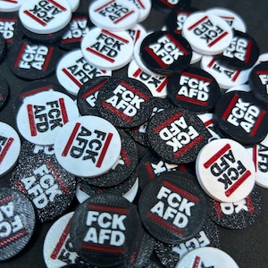 Könnte beinhalten: Eine Ansammlung kleiner, runder Spielsteine mit dem Text "FCK AFD" in fetter Schrift. Die Spielsteine sind hauptsächlich schwarz und weiß, mit roten Akzenten. Der Text ist von einem roten rechteckigen Rahmen umgeben.