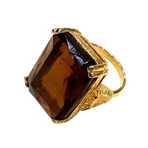 May include: A gold-toned ring featuring a large, rectangular, brown gemstone. The ring has an ornate, textured band and setting, with a classic, vintage aesthetic. The gemstone is a rich, dark brown color.