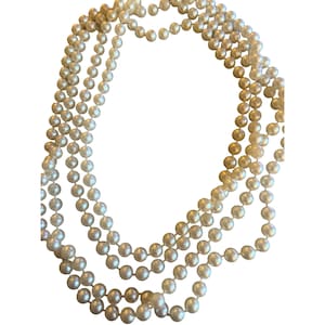 May include: A close-up of several strands of faux pearl necklaces. The pearls are a creamy white hue and are strung together. The necklaces are draped and layered, creating a classic and elegant appearance. The background is plain white.