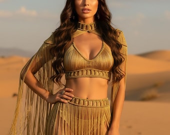 May include: A gold-colored, fringed two-piece outfit with a matching crown. The top is a halter style, and the skirt has a high slit. The outfit is worn in a desert setting.
