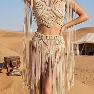 May include: A beige macrame two-piece set consisting of a high-neck cropped top and a matching floor-length skirt. Both pieces feature intricate knotted patterns and long, flowing fringe details that extend from the shoulders and waistline.