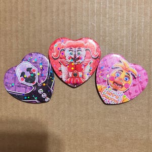May include: Three heart-shaped buttons featuring characters from the Five Nights at Freddy's franchise. One button has a purple background with a puppet design, another has a red background with a character, and the third has a pink background with a chicken character and the text "LET'S PARTY!"