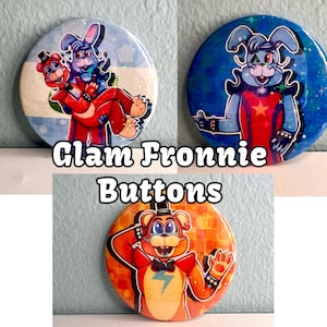 May include: Three round buttons featuring cartoon characters from the video game "Five Nights at Freddy's". The buttons are decorated with glitter and feature the characters Glamrock Freddy, Glamrock Chica, and Glamrock Monty. The text "Glam Fronic Buttons" is printed below the buttons.