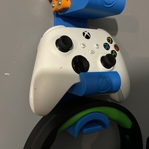 Könnte beinhalten: Blauer, an der Wand montierter Gaming-Zubehörhalter. Er verfügt über einen weißen Xbox-Controller, ein schwarzes Headset mit grünen Akzenten und einen Bereich zum Halten von orangefarbenen AA-Batterien. Der Halter hat das Xbox-Logo.