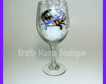 May include: A clear glass wine glass with a painted design of a crab holding the Maryland state flag. The crab is painted in blue, yellow, black, and white.