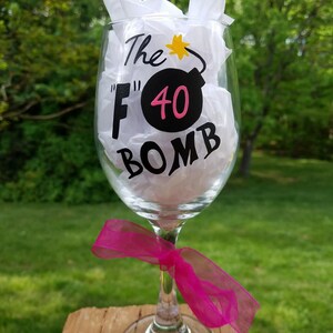 May include: A clear glass wine glass with a black and pink design that says "The "F" 40 Bomb". The glass has a pink ribbon tied around the stem.