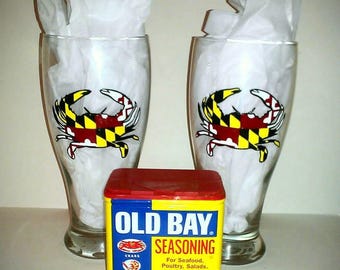 May include: Two clear glass beer mugs with a Maryland crab painted on each. A yellow and red Old Bay Seasoning tin is in the centre of the image. The tin has a label that reads "Old Bay Seasoning" and lists "For Seafood, Poultry, Salads, Meats". The label also states "Same great taste for over 60 years" and "Net Wt 6 oz 170 g".