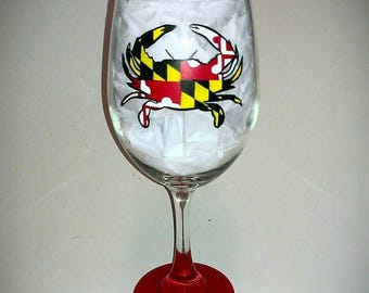 May include: A clear wine glass with a red base and a crab design. The crab is in the center of the glass and features the Maryland state flag colours. The glass is filled with white tissue paper.