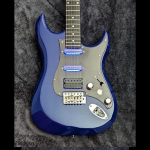 May include: A blue electric guitar with a carbon fiber pickguard and blue LED lights on the pickups. The guitar has a single cutaway body shape and a chrome bridge.
