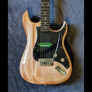 May include: A natural wood electric guitar with a black pickguard and a green pickup. The guitar has a unique body shape with a pointed headstock.