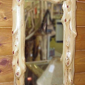 May include: Rectangular mirror with a rustic log frame. The frame is made of light-colored wood with visible knots and a natural, textured surface. The mirror reflects a blurred interior scene, adding depth to the design. A decorative piece for a cabin or rustic decor.