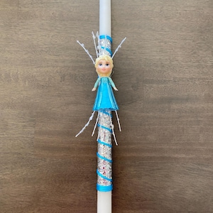 May include: A white candle with a blue and silver glitter ribbon wrapped around it. A small plastic figurine of a girl with blonde hair wearing a blue dress is attached to the top of the candle.