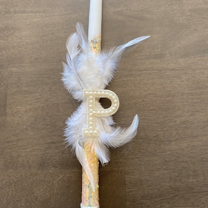 May include: White baptism candle with a gold glitter and pearl design. The candle has a white feather trim and a gold letter "P" with pearl accents.