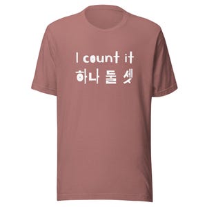 Peut inclure: T-shirt rose poussiéreux avec texte blanc. Le texte dit "I count it" en anglais, suivi de caractères coréens. Le t-shirt a des manches courtes et un col rond.